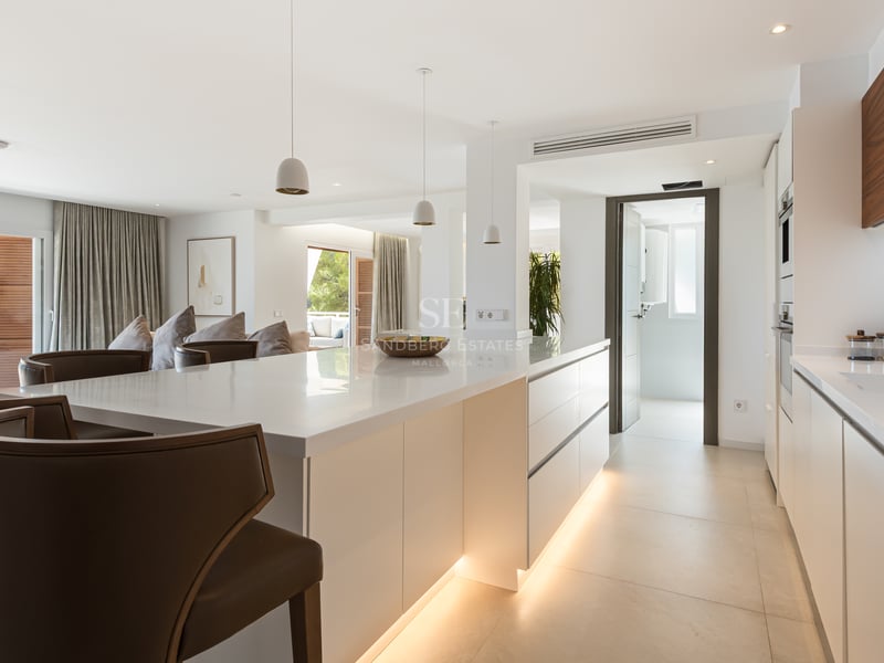Cuisine moderne lumineuse avec grand îlot blanc, tabourets en cuir et menuiserie en noyer intégrée.