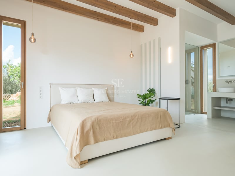 Minimalist bedroom featuring exposed wooden ceiling beams, white microcement floors, and en-suite bathroom.