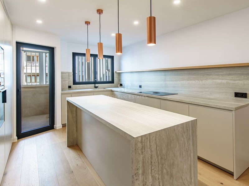 Cocina luminosa con gran isla de piedra travertino, lámparas de cobre y suelo de madera clara.