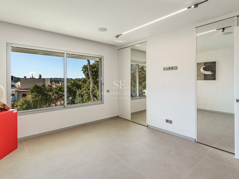 Chambre moderne avec sol gris, placards en miroir et grande fenêtre donnant sur la verdure.