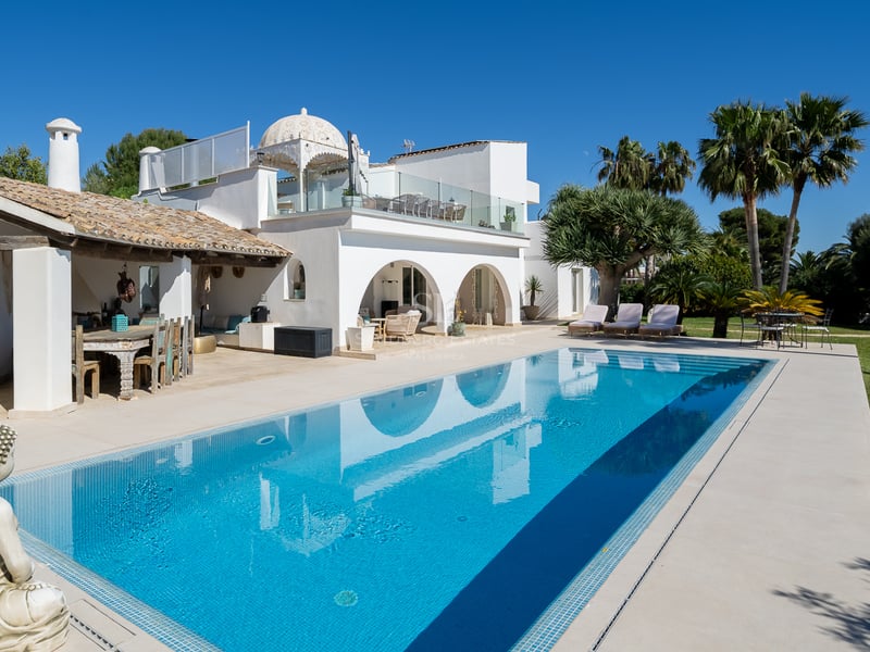 Une grande piscine bleue reflétant une villa méditerranéenne blanche avec des arches, des palmiers et une statue de Bouddha.