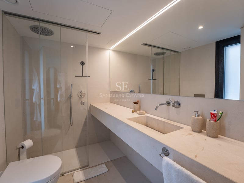 Salle de bain moderne avec vanité en pierre crème, douche à l'italienne en verre, grand miroir et éclairage linéaire.