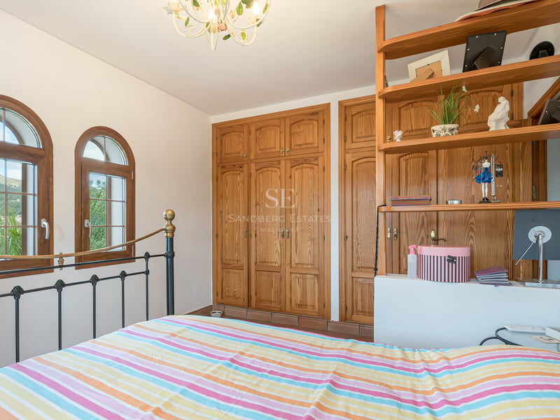 Chambre avec lit en fer forgé, armoires en bois intégrées et fenêtres cintrées offrant une vue sur le paysage.