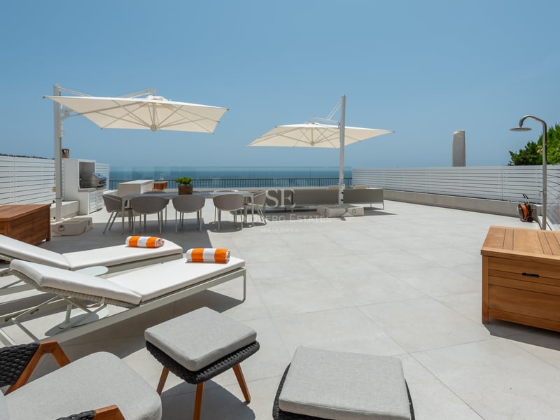 Grande terrasse blanche avec bains de soleil, coin repas, douche extérieure et garde-corps en verre face à la mer.