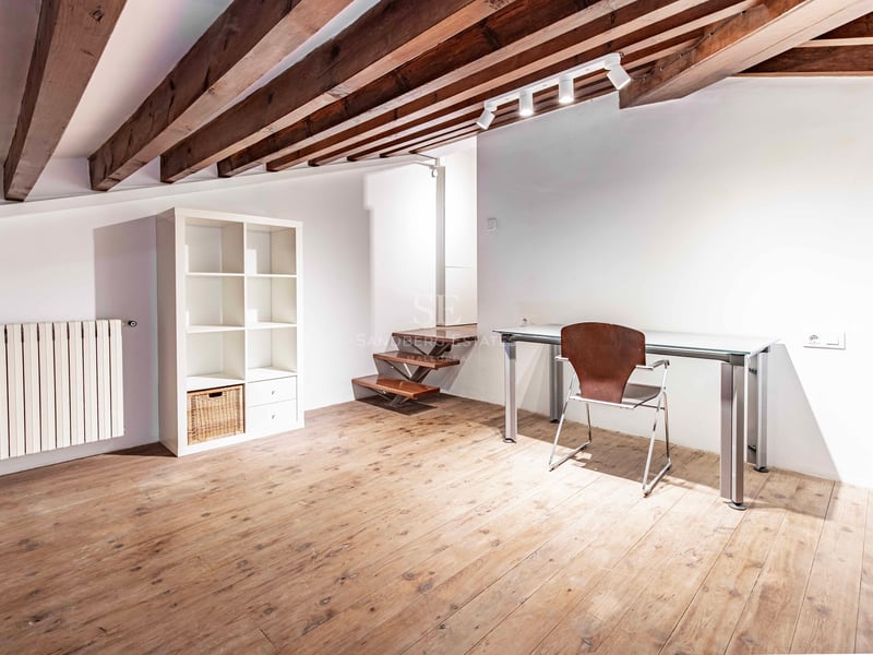 Bureau sous les toits avec poutres en bois apparentes, bureau en verre, parquet et étagère blanche minimaliste.