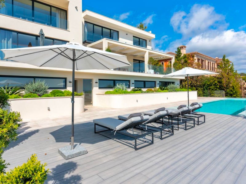 Extérieur d'une villa moderne avec piscine turquoise, chaises longues grises et parasols blancs dans un cadre paysager.