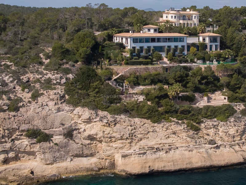 Vue aérienne d'une grande villa méditerranéenne blanche perchée sur une falaise calcaire surplombant la mer bleue.