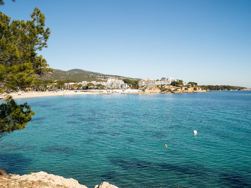 Vue panoramique d'une baie méditerranéenne aux eaux turquoises, plage de sable et bâtiments blancs sous un ciel bleu.