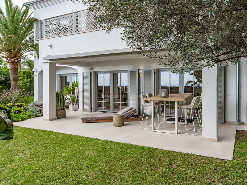 Villa méditerranéenne blanche avec patio couvert, mange-debout, chaise longue, pelouse verte et palmiers.