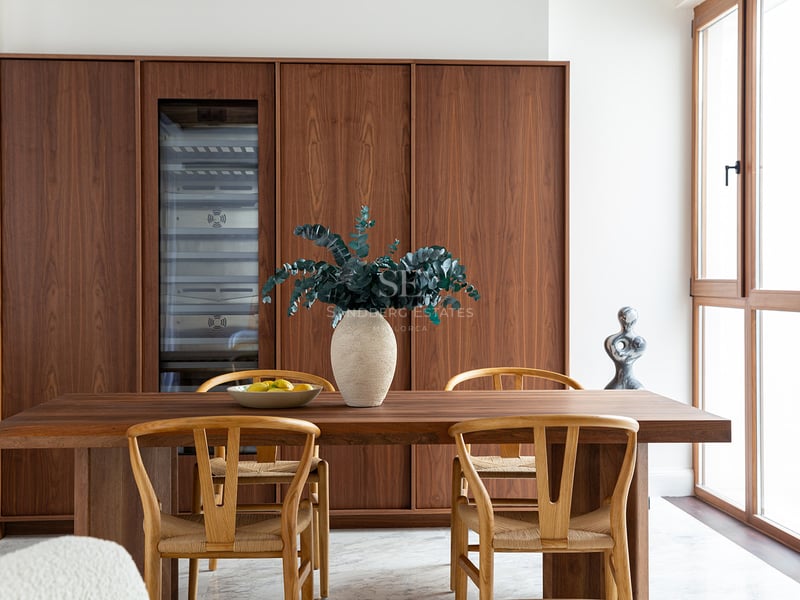 Comedor moderno con armarios de nogal, vinoteca, mesa de madera, sillas wishbone y suelos de mármol blanco.