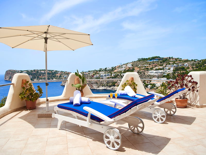 Deux bains de soleil bleus et un parasol blanc sur une terrasse en pierre avec vue panoramique sur la mer.