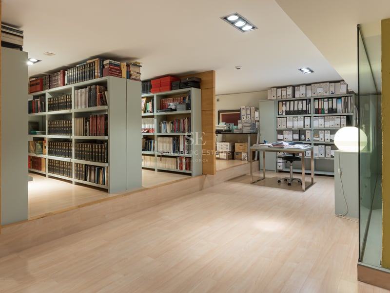 Spacious home office with extensive book shelving, light wood floors, and a modern glass-top desk.