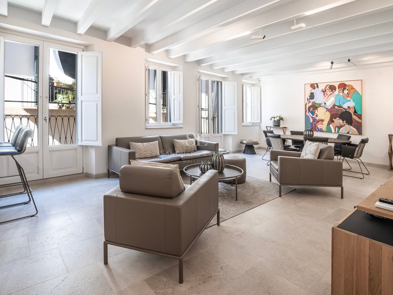 Salon lumineux avec canapés en cuir gris, plafond aux poutres blanches et coin repas avec une œuvre d'art colorée.