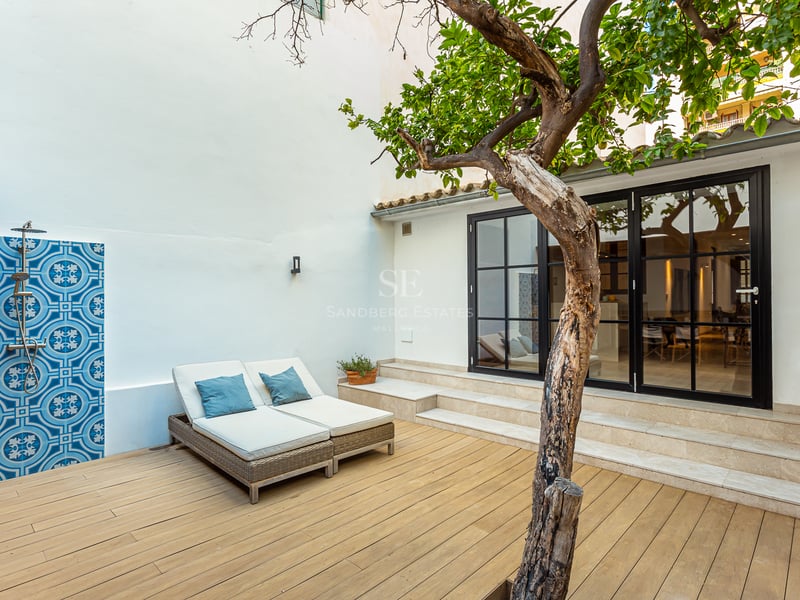 Terrasse en bois avec transats, douche extérieure aux carreaux bleus et un arbre devant de grandes baies vitrées.