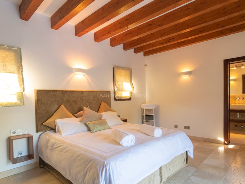 Chambre à coucher avec poutres en bois sombre, sol en pierre et salle de bain attenante.