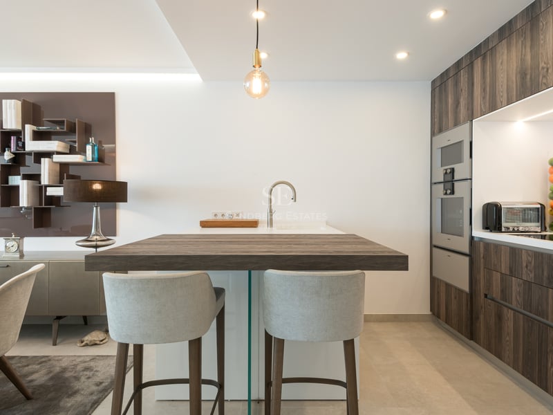 Cuisine moderne avec îlot en bois foncé, tabourets gris, armoires épurées et appareils électroménagers intégrés.