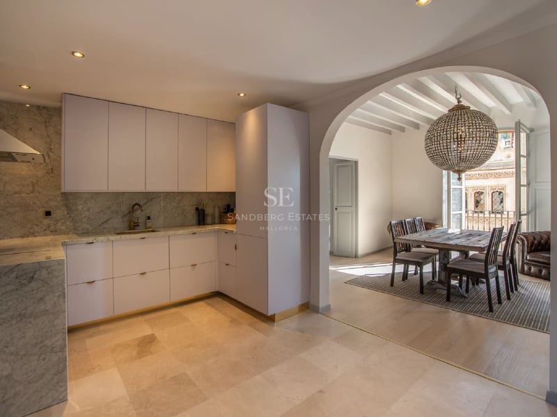 Cocina moderna blanca con encimeras de mármol que conecta mediante un arco con un comedor de madera y lámpara de cristal.