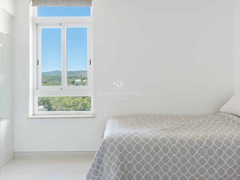 Chambre minimaliste blanche avec lit gris et fenêtre donnant sur des collines verdoyantes et un ciel bleu.
