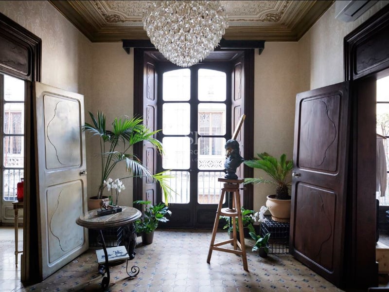 Gran entrada con puertas altas de madera, suelos de mosaico, una lámpara de cristal y plantas de interior.