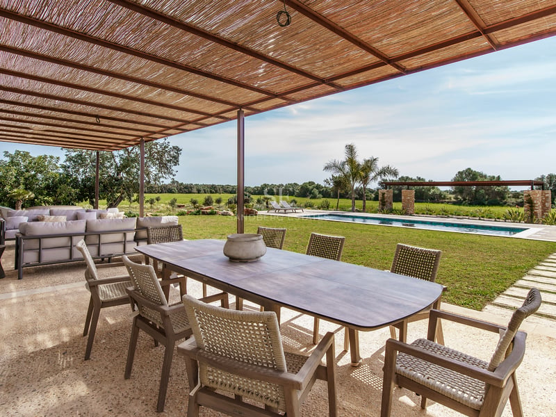Gran mesa de comedor de madera bajo pérgola de cañizo en terraza de piedra frente a una piscina y jardín.
