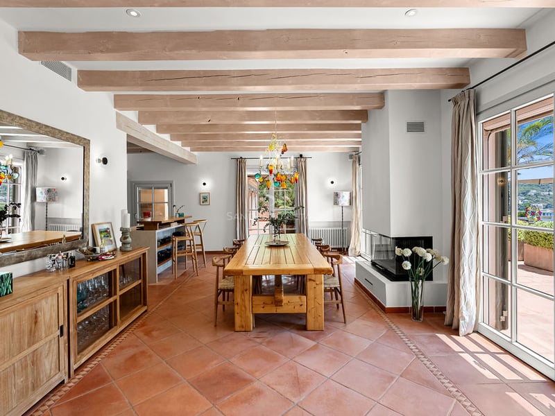 Grande salle à manger avec table en bois, sol en terre cuite et poutres au plafond menant à la terrasse.