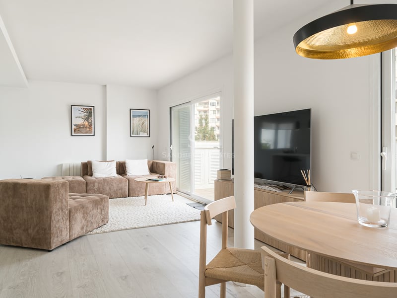 Salon ensoleillé avec un canapé modulaire marron, une table à manger en bois et de grandes baies vitrées ouvrant sur un balcon.