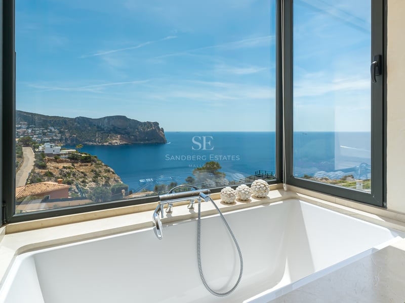 Baignoire blanche moderne devant de grandes baies vitrées donnant sur une côte rocheuse et la mer bleue.