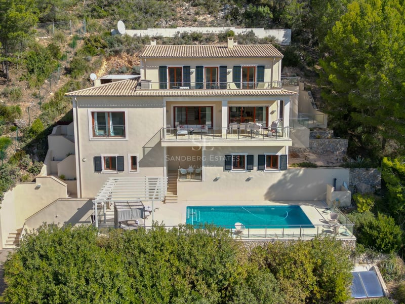 Vue aérienne d'une villa ocre de trois étages avec piscine, entourée d'une forêt de pins sur une colline.