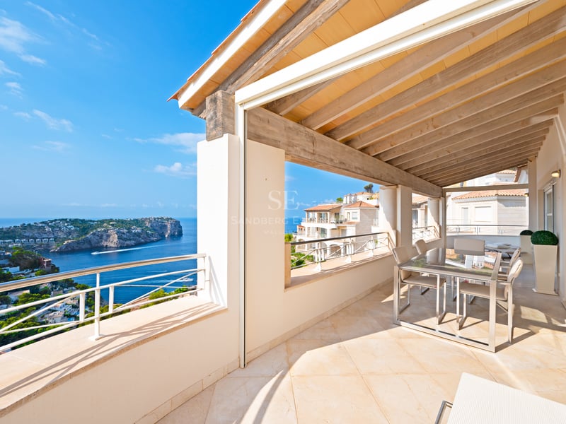 Terrasse ensoleillée avec poutres en bois et salle à manger donnant sur la mer bleue et la côte rocheuse.