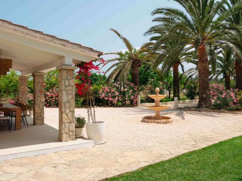 Jardin méditerranéen avec terrasse en pierre, fontaine à trois niveaux et palmiers sous un ciel bleu.