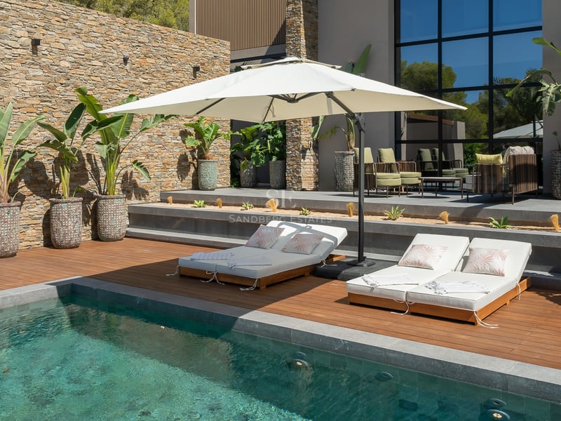 Piscine moderne avec terrasse en bois, bains de soleil blancs et parasol devant un mur en pierre naturelle.