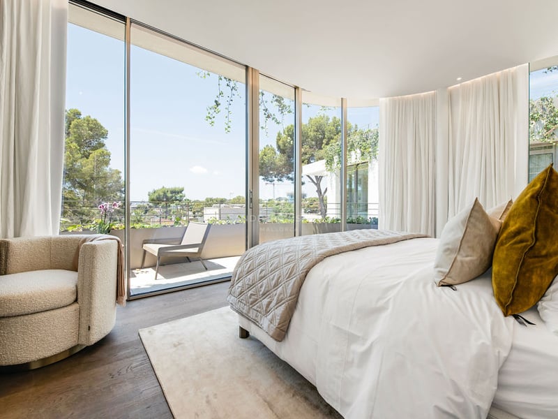 Chambre de luxe avec baies vitrées du sol au plafond, fauteuil en bouclé et vue sur une terrasse privée ensoleillée.