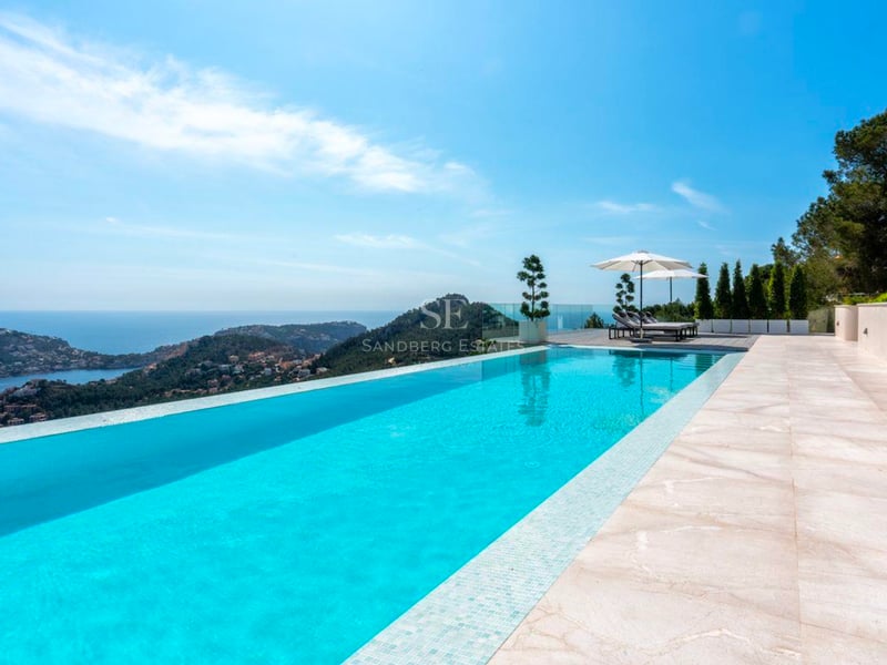 Piscine à débordement turquoise avec vue sur la mer et les montagnes, entourée de dalles en pierre et parasols blancs.