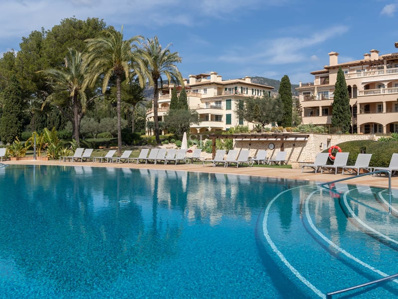 Grande piscine turquoise avec chaises longues et palmiers devant un immeuble d'appartements méditerranéen.