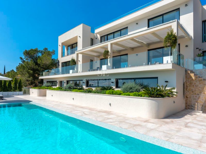 Grande piscine turquoise devant une villa blanche moderne de trois étages avec balcons en verre et escaliers en pierre.