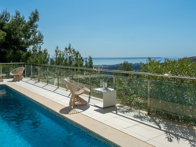Terrasse de piscine moderne avec garde-corps en verre surplombant les pins et la mer bleue sous un ciel dégagé.