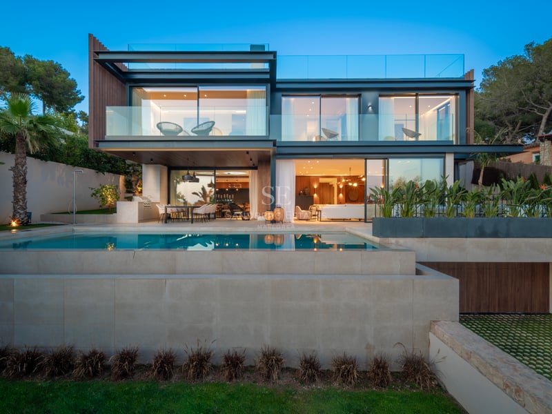 Villa moderne de deux étages avec balcons en verre et piscine à débordement éclairée au crépuscule.