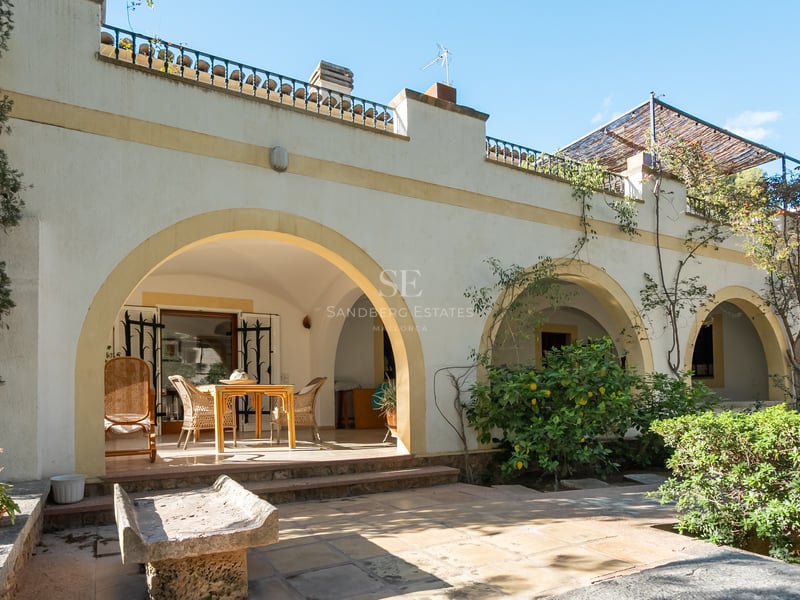 Façade de villa blanche avec de grandes arches, coin repas ombragé, banc en pierre et végétation méditerranéenne.