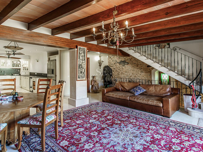 Salon ouvert avec poutres en bois sombre, mur en pierre, canapé en cuir et tapis d'orient.