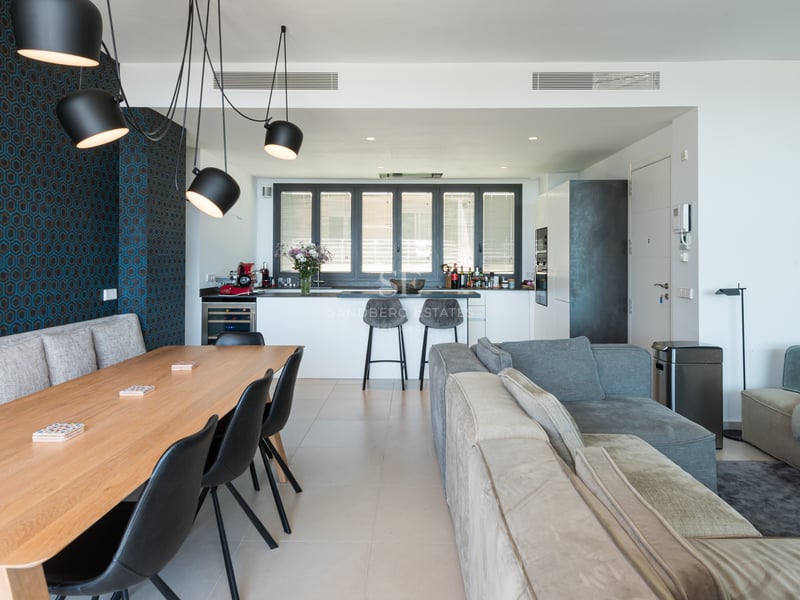 Pièce à vivre ouverte comprenant une table en bois, des canapés gris, un mur géométrique bleu et une cuisine moderne.