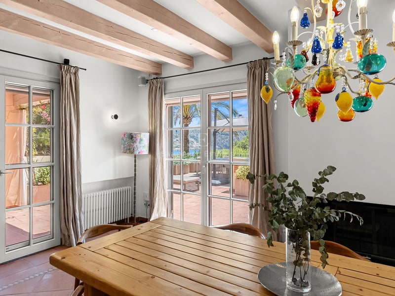 Salle à manger intérieure avec table en bois, lustre coloré, poutres apparentes et portes ouvrant sur une terrasse.