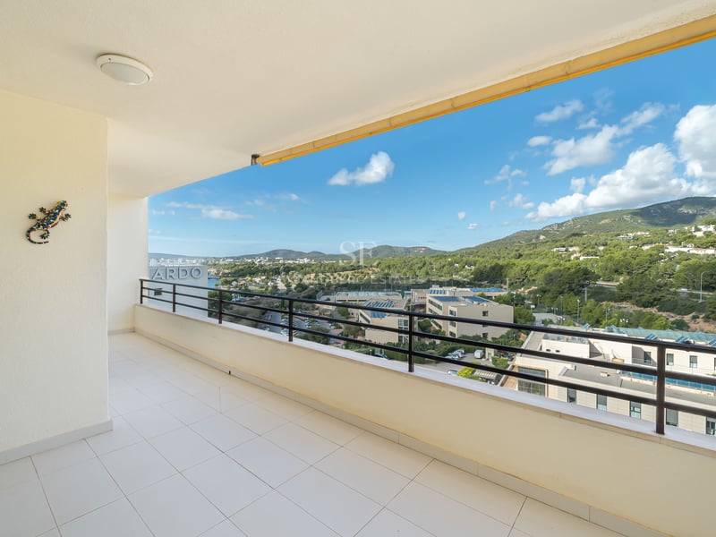 Grand balcon en carrelage blanc avec rambarde noire offrant une vue sur des collines verdoyantes et un ciel bleu.