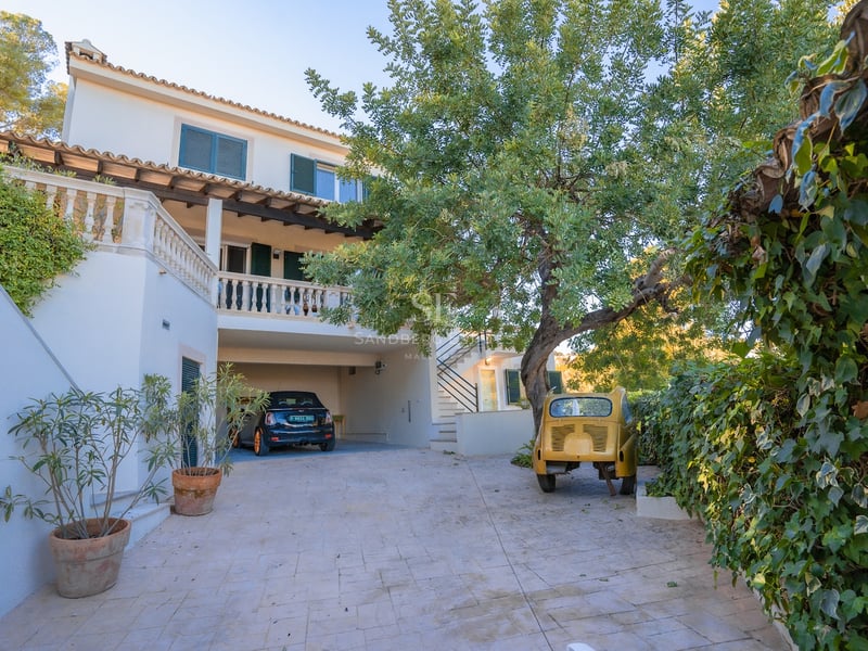 Allée pavée menant à un parking couvert dans une villa blanche avec des volets verts et des arbres.