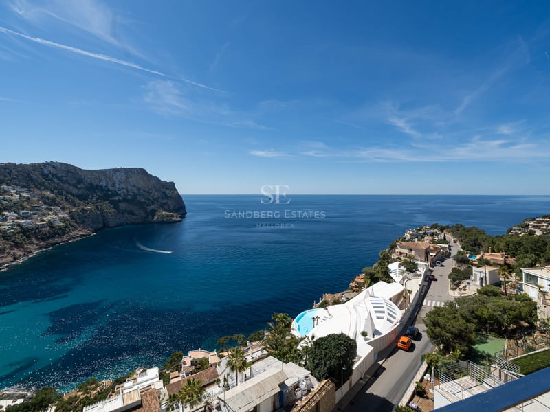 Vue en hauteur d'une baie bleu azur entourée de falaises rocheuses et de villas de luxe sous un ciel dégagé.