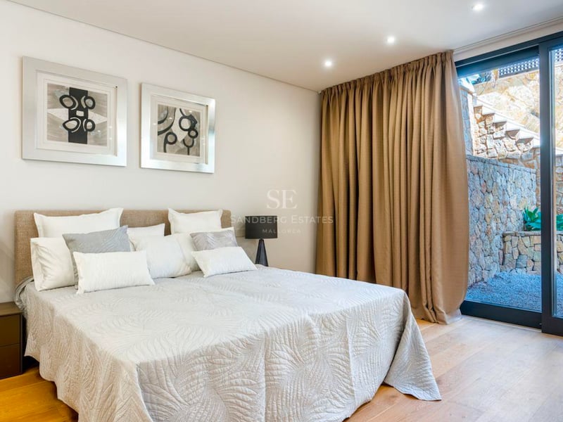 Chambre lumineuse avec lit double, tableaux abstraits et grandes baies vitrées ouvrant sur un patio en pierre.