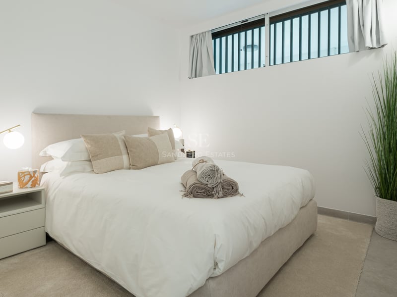 Chambre lumineuse avec lit rembourré beige, linge blanc, lampes en laiton et plante d'intérieur haute.