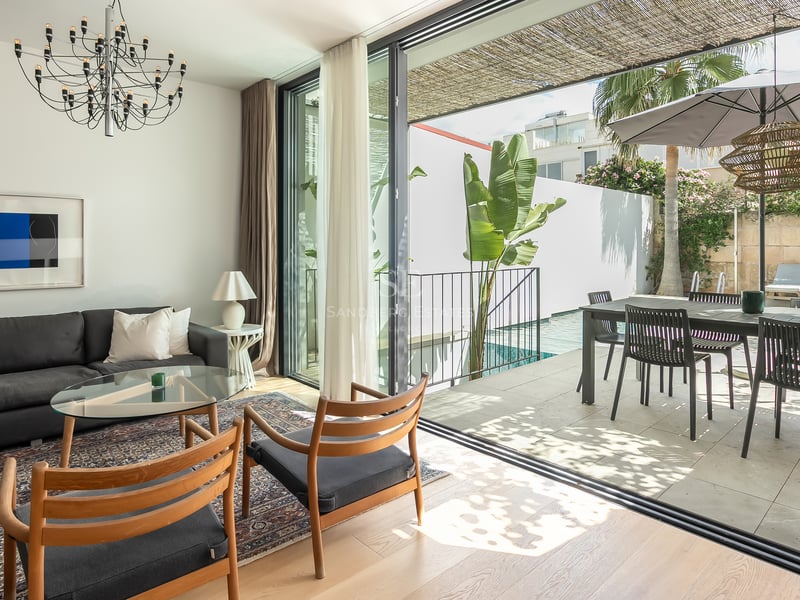 Salon lumineux avec canapé sombre et chaises en bois ouvrant sur une terrasse ensoleillée avec piscine et coin repas.
