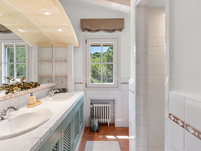 Salle de bain lumineuse avec deux lavabos blancs, meubles verts, sol en terre cuite et vue sur les arbres.