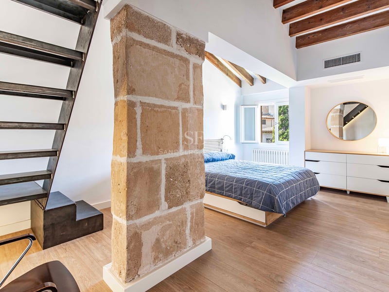 Chambre lumineuse avec pilier en pierre naturelle, escalier en acier industriel, poutres en bois et lit double.