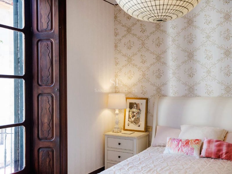 Dormitorio con papel tapiz estampado, cama blanca, lámpara de noche y marco de ventana de madera oscura.